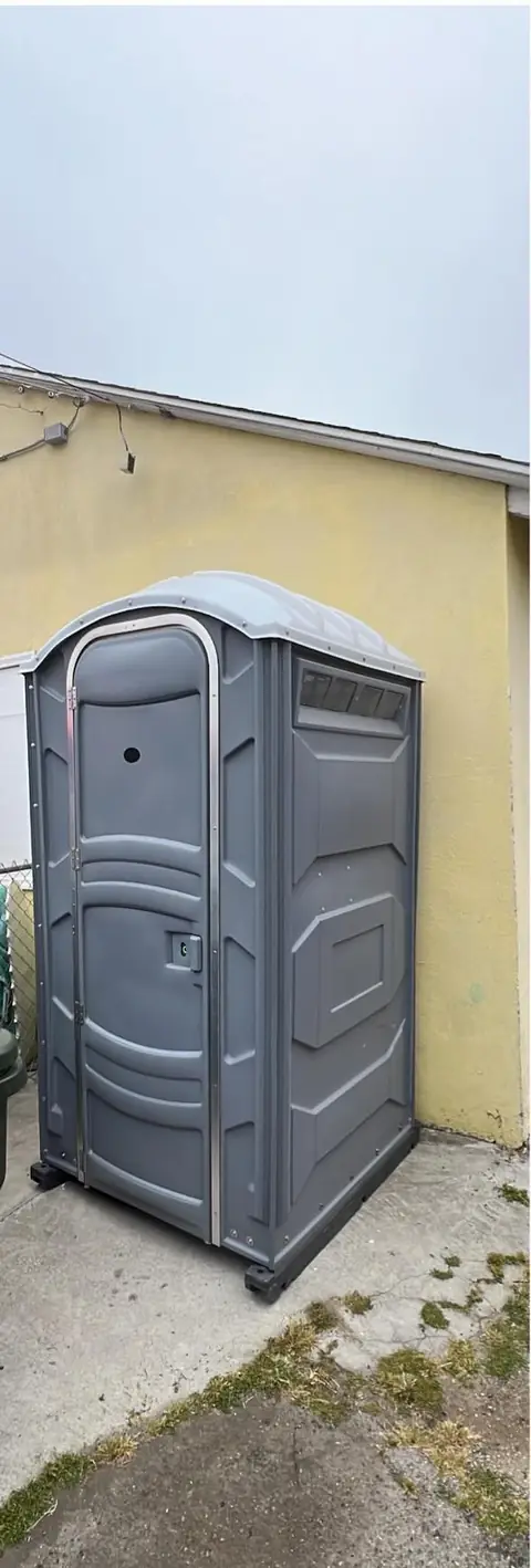 Gray porta potty in a narrow side yard next to a house